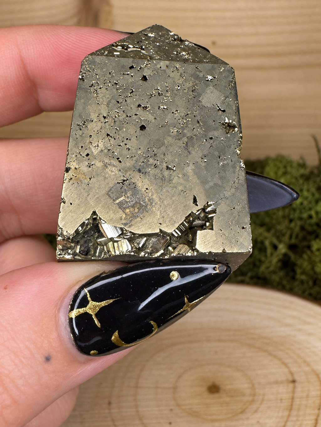 A hand holding a raw pyrite crystal cluster with metallic gold and gray-brown mineral texture, shown close-up against a wood and moss background.